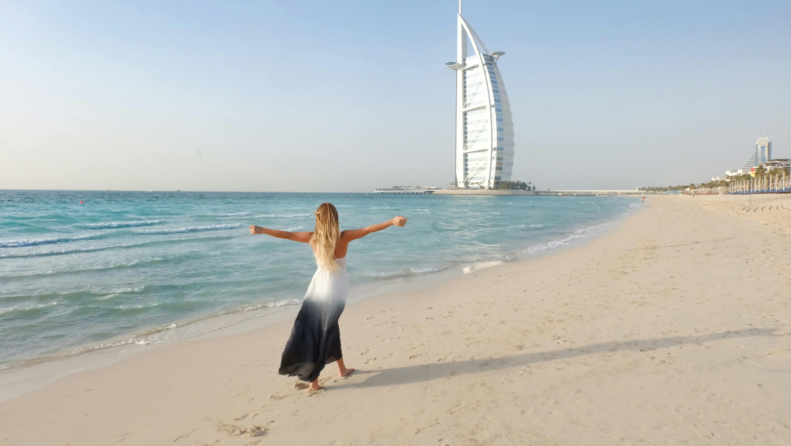 A carefree woman in a flowing dress strolls along the Dubai beach with Burj Al Arab in view.